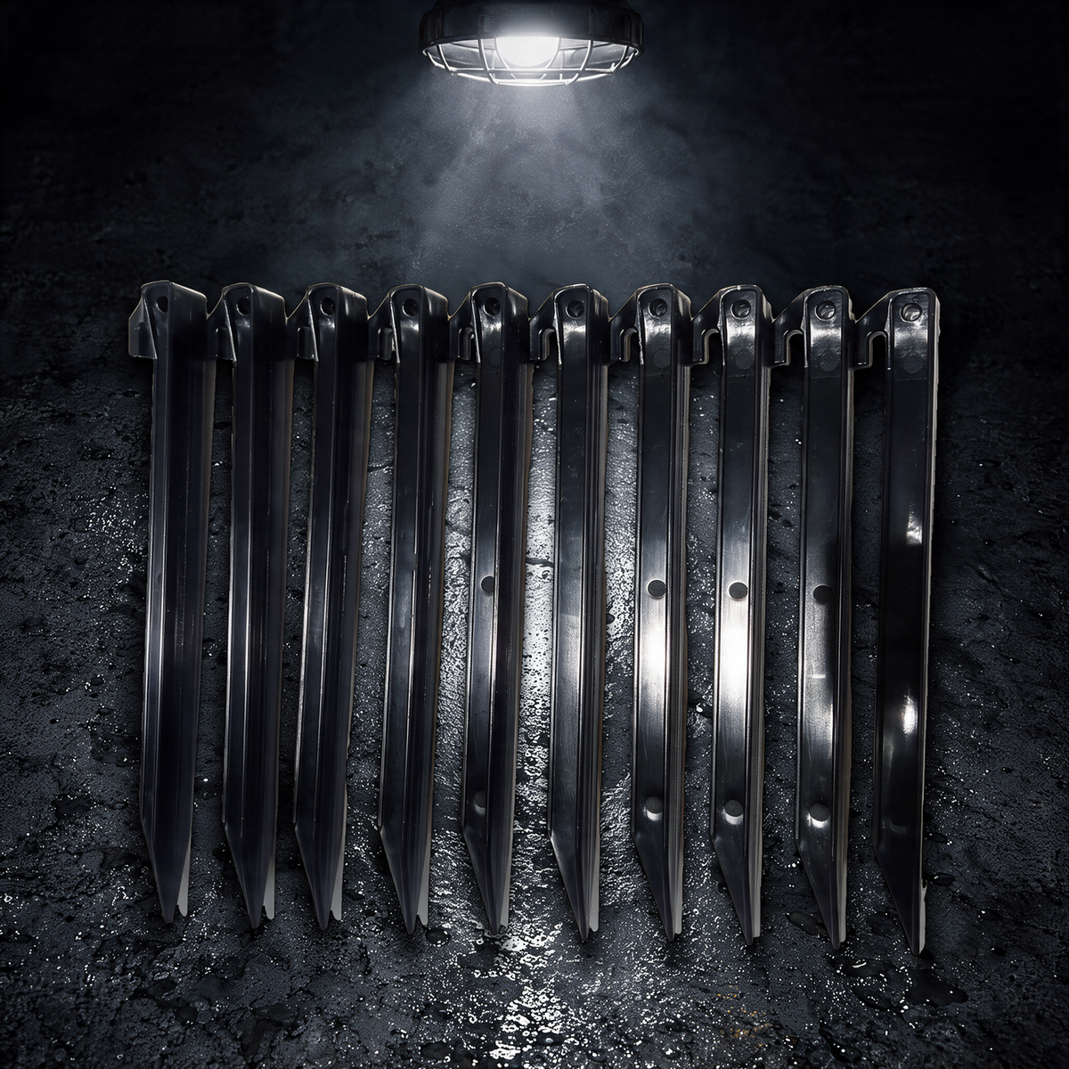 Ten black BruteLine 300mm ground pegs lying flat in a straight row on wet concrete under a work lamp, each with a line hook and tie-off hole