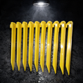 Ten yellow BruteLine 300mm ground pegs lying flat in a straight row on wet concrete under a work lamp, each with a line hook and tie-off hole