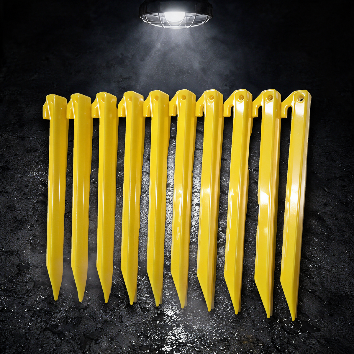 Ten yellow BruteLine 300mm ground pegs lying flat in a straight row on wet concrete under a work lamp, each with a line hook and tie-off hole